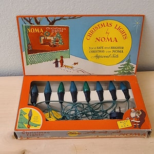 May include: A box of vintage NOMA Christmas lights with a green cord and eight green and blue bulbs. The box has a picture of a winter scene with children and a dog. The box also has the text "Christmas Lights by NOMA" and "For a safe and brighter Christmas use NOMA Approved Sets".