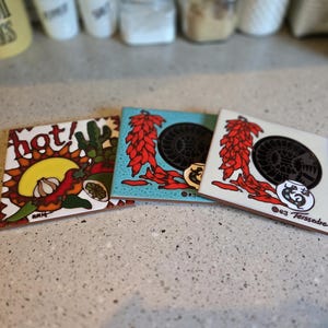 May include: Three ceramic coasters with colorful designs. One coaster features a sun, chili peppers, garlic, and the word "hot!" Another coaster has a blue background with red chili peppers and a black and white design. The third coaster has a white background with red chili peppers and a black and white design.