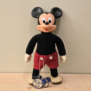 Peut inclure: Une poup&eacute;e en peluche de Mickey Mouse, avec un col roul&eacute; noir, un short rouge et de grandes oreilles noires. La poup&eacute;e a des gants et des chaussures blancs, avec une &eacute;tiquette "Guided Tour".