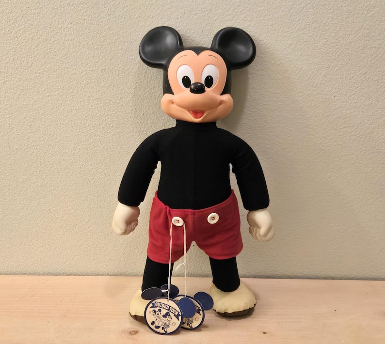 Peut inclure: Une poup&eacute;e en peluche de Mickey Mouse, avec un col roul&eacute; noir, un short rouge et de grandes oreilles noires. La poup&eacute;e a des gants et des chaussures blancs, avec une &eacute;tiquette "Guided Tour".