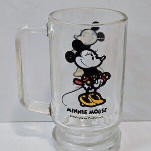 May include: A clear glass mug featuring a graphic of Minnie Mouse wearing a red and yellow polka dot dress and yellow shoes. The text MINNIE MOUSE is printed in black below the character, followed by the text &copy;Walt Disney Productions.