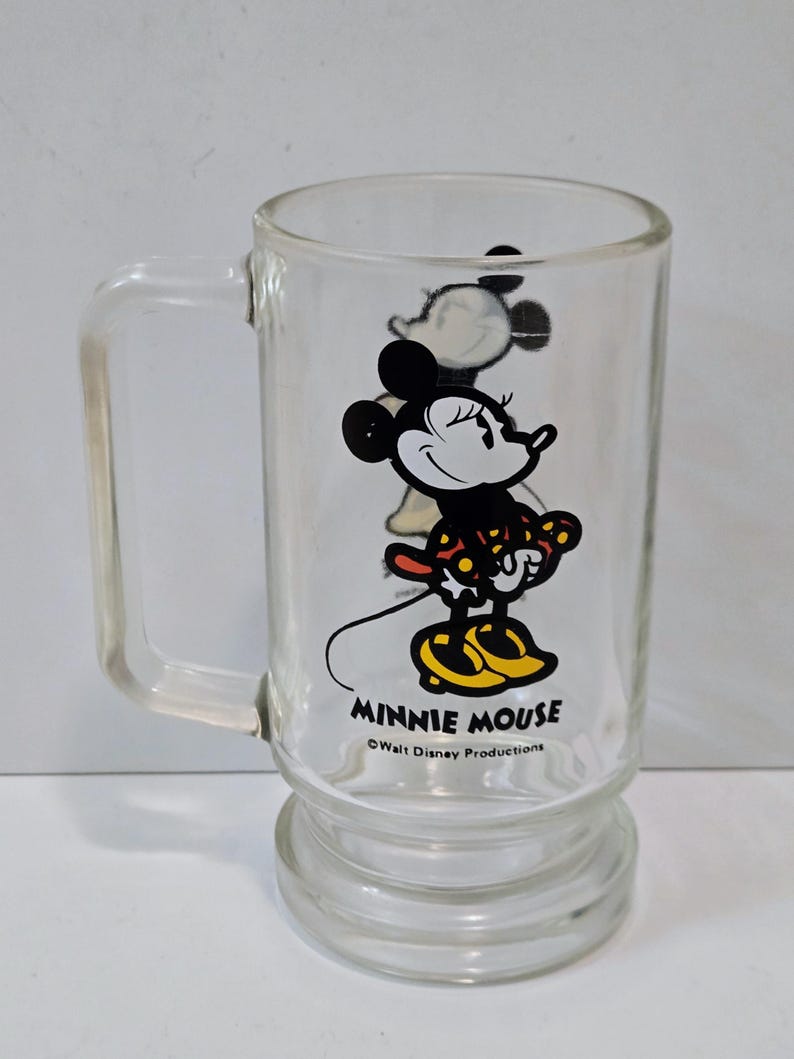 May include: A clear glass mug featuring a graphic of Minnie Mouse wearing a red and yellow polka dot dress and yellow shoes. The text MINNIE MOUSE is printed in black below the character, followed by the text &copy;Walt Disney Productions.