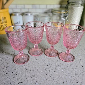 May include: Four pink vintage goblets with a detailed starburst pattern. The glasses have a stem and a rounded bowl. The goblets are arranged on a speckled countertop, suggesting a kitchen or dining setting.