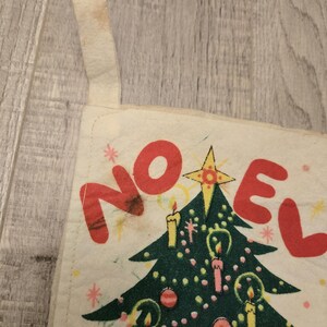 May include: A vintage Christmas stocking with a green Christmas tree and the word "NOEL" in red letters. The stocking is made of white fabric with a red and white trim.