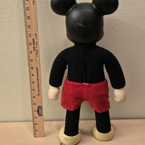 Peut inclure: Une poup&eacute;e en peluche vintage de Mickey Mouse, vue de dos. La poup&eacute;e a une t&ecirc;te et des oreilles noires, un corps noir, un short rouge et des mains et des pieds cr&egrave;me. Une r&egrave;gle en bois est plac&eacute;e &agrave; c&ocirc;t&eacute; de la poup&eacute;e pour l'&eacute;chelle.