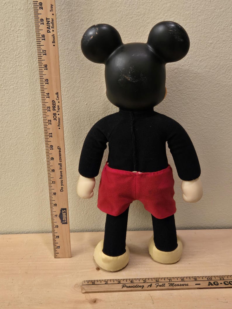 Peut inclure: Une poup&eacute;e en peluche vintage de Mickey Mouse, vue de dos. La poup&eacute;e a une t&ecirc;te et des oreilles noires, un corps noir, un short rouge et des mains et des pieds cr&egrave;me. Une r&egrave;gle en bois est plac&eacute;e &agrave; c&ocirc;t&eacute; de la poup&eacute;e pour l'&eacute;chelle.