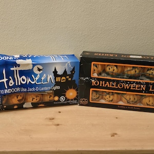 May include: Two boxes of Halloween lights. One box is blue with the word "Halloween" and a silhouette of a castle and a bat. The other box is black and has the words "10 Halloween Lights". Both boxes contain Jack-O-Lantern lights.