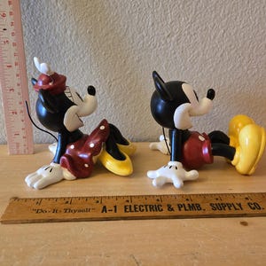 Puede incluir: Dos figuras de cer&aacute;mica de Mickey y Minnie Mouse. Minnie lleva un vestido rojo con lunares blancos, un lazo a juego y zapatos amarillos. Mickey lleva pantalones cortos rojos y zapatos amarillos. Ambos son negros, blancos y rojos.