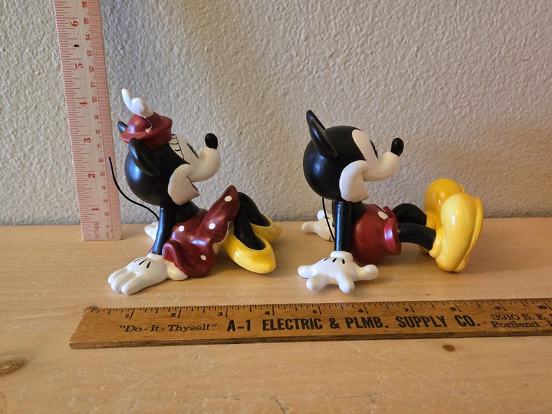 Puede incluir: Dos figuras de cer&aacute;mica de Mickey y Minnie Mouse. Minnie lleva un vestido rojo con lunares blancos, un lazo a juego y zapatos amarillos. Mickey lleva pantalones cortos rojos y zapatos amarillos. Ambos son negros, blancos y rojos.