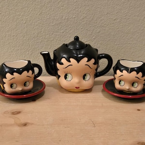 May include: A vintage ceramic tea set featuring a black teapot and two matching teacups. The teapot and cups are shaped like a cartoon character's face, with black hair, large eyes, and red lips. The cups sit on black saucers with red rims.