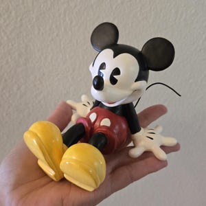 Puede incluir: Una figura de Mickey Mouse, con cabeza negra y orejas grandes, cara blanca y nariz negra. La figura lleva cuerpo negro, pantalones cortos rojos, zapatos amarillos y guantes blancos. Un objeto de colecci&oacute;n cl&aacute;sico de Disney.
