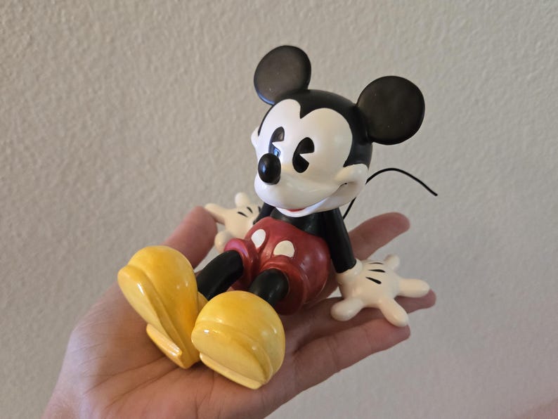 Puede incluir: Una figura de Mickey Mouse, con cabeza negra y orejas grandes, cara blanca y nariz negra. La figura lleva cuerpo negro, pantalones cortos rojos, zapatos amarillos y guantes blancos. Un objeto de colecci&oacute;n cl&aacute;sico de Disney.