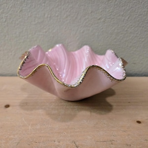 May include: A pink, clam-shaped ceramic bowl with a wavy rim and gold trim. The bowl has a glossy finish and sits on a wooden surface. The interior has a swirled pattern. A decorative piece for serving or display.