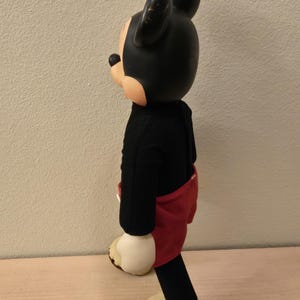 Peut inclure: Poup&eacute;e vintage de Mickey Mouse, avec une t&ecirc;te et des oreilles noires, un corps noir, un short rouge et des mains et pieds cr&egrave;me. La poup&eacute;e est pos&eacute;e sur une surface en bois.