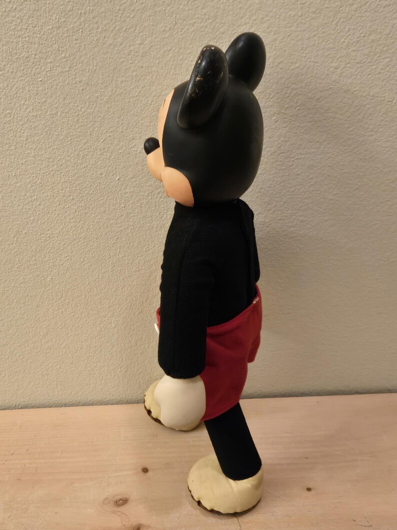 Peut inclure: Poup&eacute;e vintage de Mickey Mouse, avec une t&ecirc;te et des oreilles noires, un corps noir, un short rouge et des mains et pieds cr&egrave;me. La poup&eacute;e est pos&eacute;e sur une surface en bois.