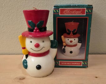 May include: A snowman candle with a red top hat, scarf, and a yellow broom. The candle is white with a black button and a red nose. A box with a similar snowman candle is next to it. The box reads "Christmas Around the World."