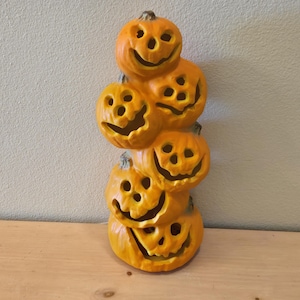 May include: A stack of six orange ceramic jack-o'-lanterns with carved smiling faces. The Halloween decoration is displayed on a wooden surface against a neutral wall. Each pumpkin has a stem.