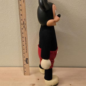 Peut inclure: Une poup&eacute;e en peluche vintage de Mickey Mouse, avec une t&ecirc;te et des oreilles noires, un pull noir, un short rouge et de grandes chaussures jaunes. La poup&eacute;e est debout, pr&eacute;sentant son design classique et son apparence embl&eacute;matique.