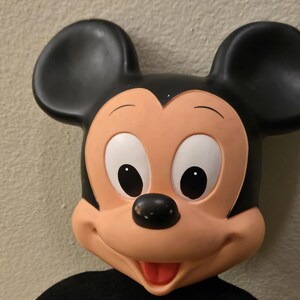 Peut inclure: Gros plan sur une figurine de Mickey Mouse. La figurine a de grandes oreilles noires, un visage couleur p&ecirc;che et un nez noir. Les yeux sont blancs avec des pupilles noires. La bouche est ouverte, r&eacute;v&eacute;lant une langue rouge. La figurine porte une chemise noire.