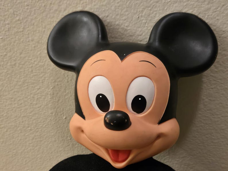 Peut inclure: Gros plan sur une figurine de Mickey Mouse. La figurine a de grandes oreilles noires, un visage couleur p&ecirc;che et un nez noir. Les yeux sont blancs avec des pupilles noires. La bouche est ouverte, r&eacute;v&eacute;lant une langue rouge. La figurine porte une chemise noire.