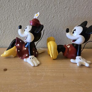 Puede incluir: Dos figuras de Mickey y Minnie Mouse en una pose relajada. Minnie lleva un vestido rojo con lunares blancos y un sombrero a juego. Mickey lleva pantalones cortos rojos. Ambos tienen guantes blancos y zapatos amarillos. Las figuras son negras, blancas, rojas y amarillas.