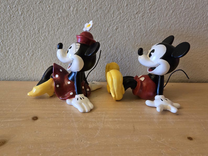 Puede incluir: Dos figuras de Mickey y Minnie Mouse en una pose relajada. Minnie lleva un vestido rojo con lunares blancos y un sombrero a juego. Mickey lleva pantalones cortos rojos. Ambos tienen guantes blancos y zapatos amarillos. Las figuras son negras, blancas, rojas y amarillas.