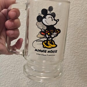 May include: A clear glass mug featuring a graphic of Minnie Mouse wearing a red and yellow polka dot dress and yellow shoes. The text MINNIE MOUSE is printed in black below the character, followed by the text Walt Disney Productions.