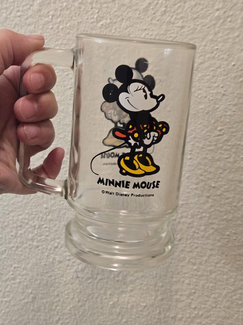 May include: A clear glass mug featuring a graphic of Minnie Mouse wearing a red and yellow polka dot dress and yellow shoes. The text MINNIE MOUSE is printed in black below the character, followed by the text Walt Disney Productions.