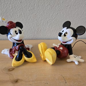 Puede incluir: Dos figuras de cer&aacute;mica de Mickey y Minnie Mouse. Mickey lleva pantalones cortos rojos y Minnie una falda y sombrero rojos. Ambos tienen orejas negras, guantes blancos y zapatos amarillos. Est&aacute;n sentados.