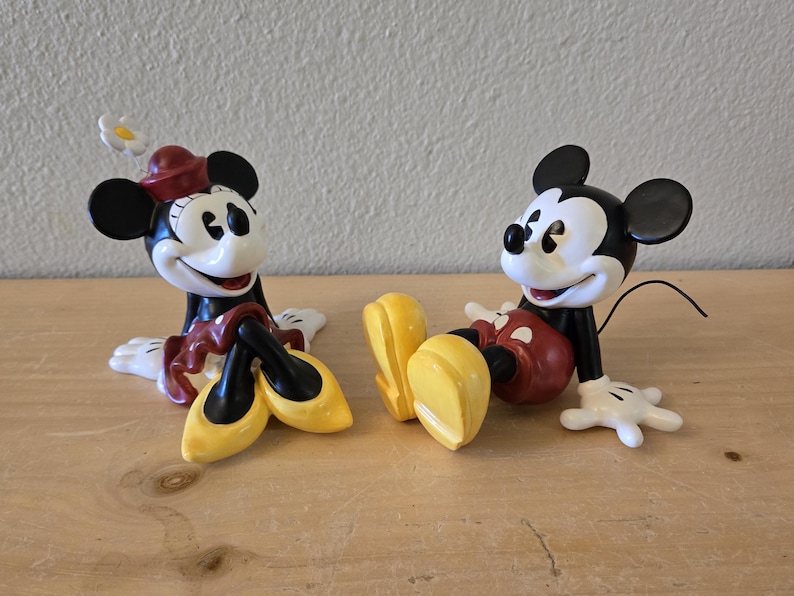 Puede incluir: Dos figuras de cer&aacute;mica de Mickey y Minnie Mouse. Mickey lleva pantalones cortos rojos y Minnie una falda y sombrero rojos. Ambos tienen orejas negras, guantes blancos y zapatos amarillos. Est&aacute;n sentados.