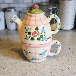May include: A decorative ceramic teapot and teacup set. The teapot is white with a pink plaid pattern, a pink rose, and blue floral accents. The teacup is stacked beneath the teapot and has a matching design.