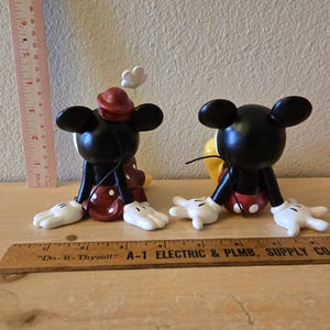 Puede incluir: Dos figuras de Mickey Mouse. Las figuras negras tienen guantes y pies blancos, pantalones cortos rojos con lunares blancos y un sombrero rojo. Uno tiene una flor blanca en la cabeza. Una regla est&aacute; al lado de las figuras.