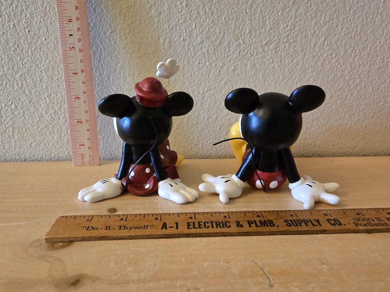Puede incluir: Dos figuras de Mickey Mouse. Las figuras negras tienen guantes y pies blancos, pantalones cortos rojos con lunares blancos y un sombrero rojo. Uno tiene una flor blanca en la cabeza. Una regla est&aacute; al lado de las figuras.