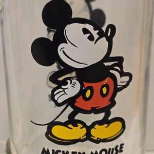 May include: A clear glass mug featuring a graphic of Mickey Mouse wearing red shorts and yellow shoes. The character is standing with hands on hips. Below the graphic, the text reads MICKEY MOUSE and &copy;Walt Disney Productions in black.