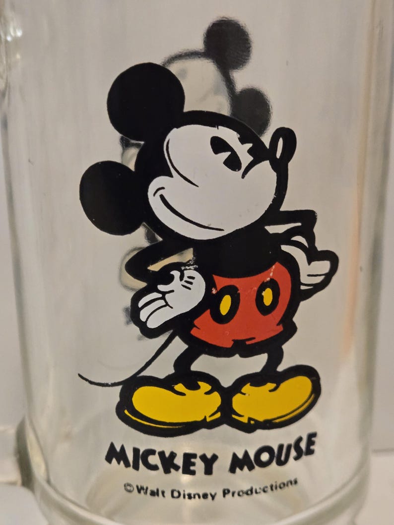 May include: A clear glass mug featuring a graphic of Mickey Mouse wearing red shorts and yellow shoes. The character is standing with hands on hips. Below the graphic, the text reads MICKEY MOUSE and &copy;Walt Disney Productions in black.