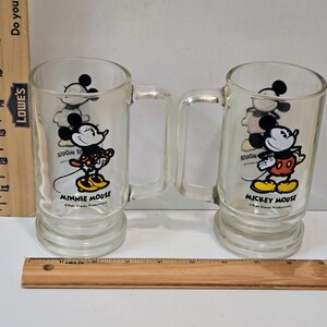 May include: Two clear glass mugs with handles, one featuring Minnie Mouse and the other Mickey Mouse. Each mug displays the character name and Walt Disney Productions text. A wooden ruler is visible, showing the mugs are approximately 6 inches tall.