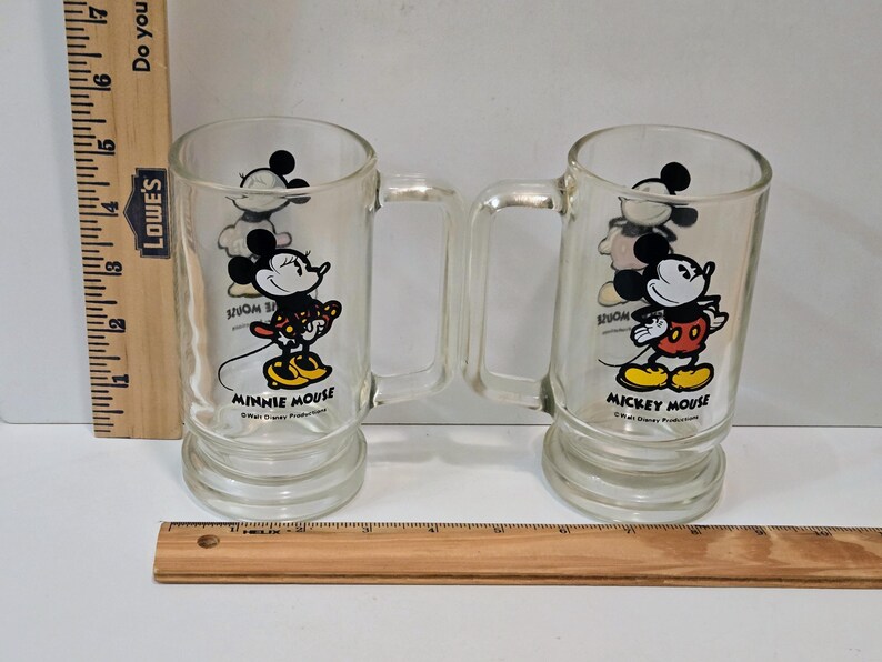 May include: Two clear glass mugs with handles, one featuring Minnie Mouse and the other Mickey Mouse. Each mug displays the character name and Walt Disney Productions text. A wooden ruler is visible, showing the mugs are approximately 6 inches tall.