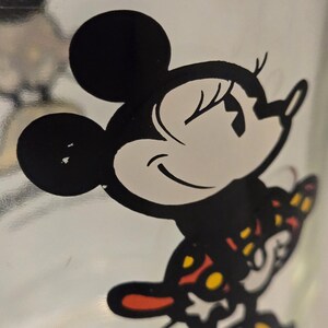 May include: A clear glass surface featuring a black and white graphic illustration of a cartoon mouse character with large round ears. The character is wearing a patterned outfit with red and yellow accents, depicted in a side profile view.