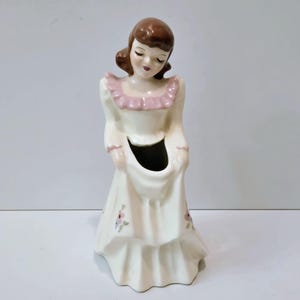 May include: A vintage ceramic figurine of a woman in a long cream-colored dress with a pink ruffled collar. The figurine has brown hair and closed eyes, with small floral accents on the dress. The figurine is a vase.