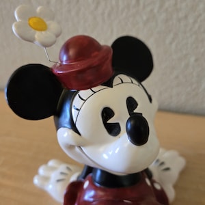 Puede incluir: Figura de Minnie Mouse con sombrero y vestido rojos con lunares blancos. La figura tiene orejas negras y cara blanca. Una flor blanca con centro amarillo est&aacute; sujeta al sombrero.