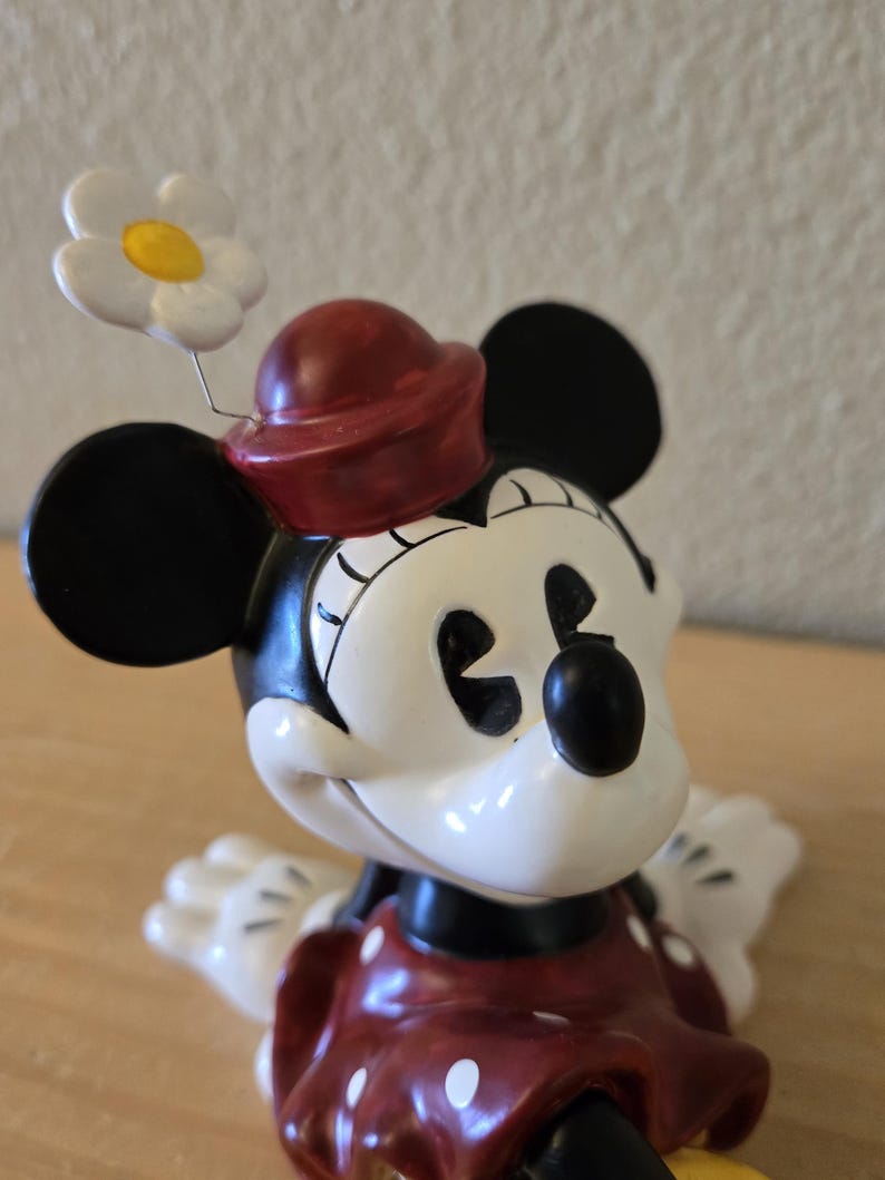 Puede incluir: Figura de Minnie Mouse con sombrero y vestido rojos con lunares blancos. La figura tiene orejas negras y cara blanca. Una flor blanca con centro amarillo est&aacute; sujeta al sombrero.