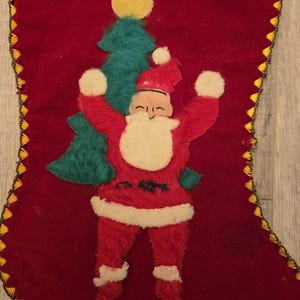 May include: A vintage red velvet Christmas stocking with a felt appliqu&eacute; of Santa Claus holding a green Christmas tree. The stocking has a yellow and gold trim.