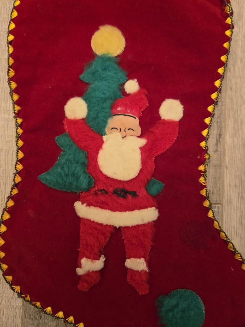 May include: A vintage red velvet Christmas stocking with a felt appliqu&eacute; of Santa Claus holding a green Christmas tree. The stocking has a yellow and gold trim.