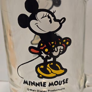 May include: A clear glass mug featuring a graphic of Minnie Mouse wearing a red dress with yellow polka dots and yellow shoes. The text MINNIE MOUSE is printed in black below the character, followed by the text &copy;Walt Disney Productions.