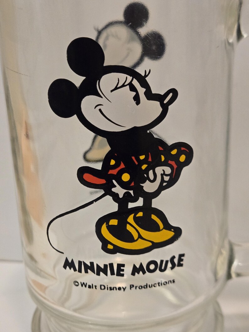 May include: A clear glass mug featuring a graphic of Minnie Mouse wearing a red dress with yellow polka dots and yellow shoes. The text MINNIE MOUSE is printed in black below the character, followed by the text &copy;Walt Disney Productions.