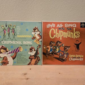 May include: Two vintage vinyl record albums featuring The Chipmunks. One album cover is teal with cartoon chipmunks and the text "The Chipmunk Song". The other album cover is orange with the text "Let's All Sing With The Chipmunks".