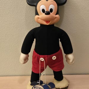 Peut inclure: Une poup&eacute;e en peluche vintage de Mickey Mouse avec des oreilles noires, un haut noir, un short rouge et des gants et chaussures blancs. La poup&eacute;e a un visage souriant et tient deux &eacute;tiquettes "Guided Tour".