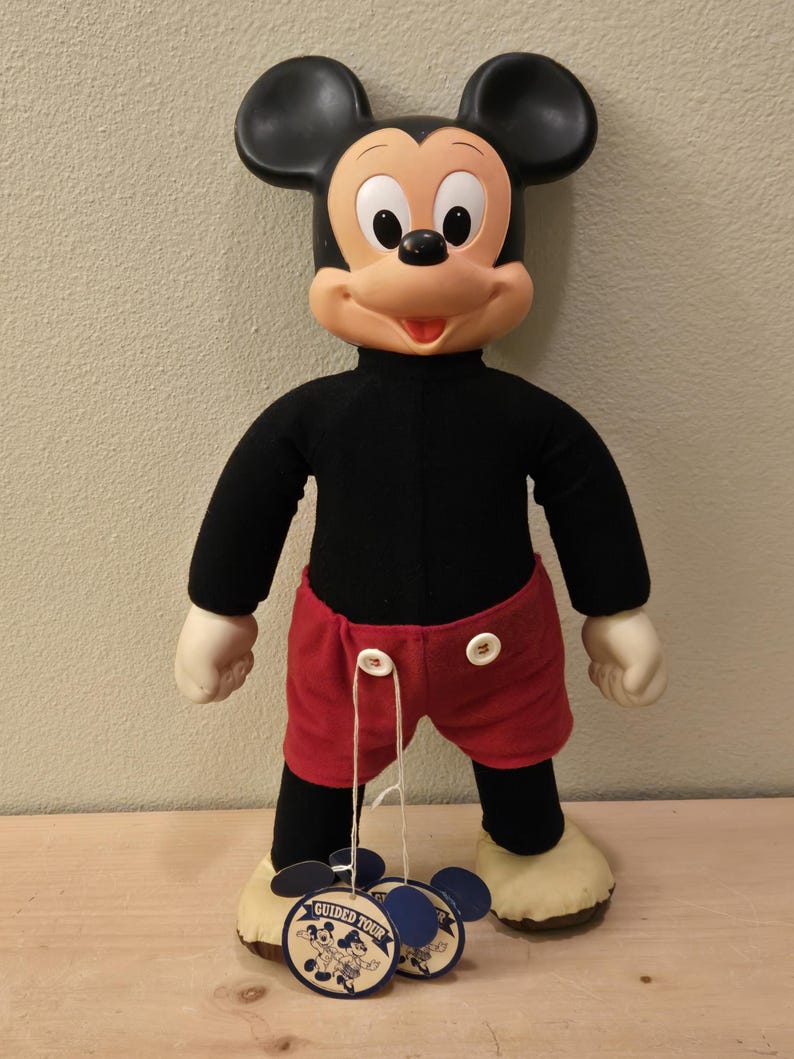Peut inclure: Une poup&eacute;e en peluche vintage de Mickey Mouse avec des oreilles noires, un haut noir, un short rouge et des gants et chaussures blancs. La poup&eacute;e a un visage souriant et tient deux &eacute;tiquettes "Guided Tour".