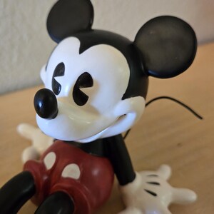 Puede incluir: Figura de Mickey Mouse con cabeza y orejas negras, cara blanca y pantalones cortos rojos. La figura tiene guantes blancos y piernas y brazos negros. Un personaje cl&aacute;sico de Disney.