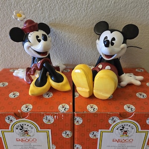 Puede incluir: Dos figuras de cer&aacute;mica de Mickey y Minnie Mouse sentadas en cajas. Minnie lleva un sombrero rojo y zapatos amarillos, y Mickey tiene zapatos amarillos. Las cajas son naranjas con dise&ntilde;os de Mickey Mouse y el logo de Enesco.
